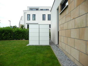 . The image depicts a modern residential area. There is a white cube-shaped structure in the foreground, which appears to be an outdoor storage unit or utility box. In the background, there is a multi-story building with large windows. The foreground includes a grassy lawn and a hedge. To the right, there's a building with a facade of large stone tiles.