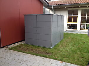 . The image shows a gray rectangular structure that appears to be a small shed or storage unit situated on a patch of grass. It is located beside a red building and is near a window with people visible inside the adjacent structure. The area also includes a pathway of paving stones.