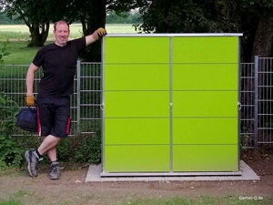 . Ein Mann lehnt lächelnd an einem grünem Gartenschrank, hält eine Werkzeugtasche, trägt Handschuhe.