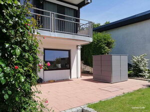 . Ein moderner Garten mit Terrasse, Pflanzen, einem Balkon und einem großen, grauen Metallschuppen.