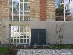 . The image shows the exterior of a building with a large grid of windows. Below the windows, there's a utility cabinet or storage unit against the wall. The wall is made of brick and concrete, and there's a partially cobbled path in front with grass and bare shrubs.