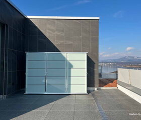 Eine moderne Dachterrasse mit Glaszaun, Blick auf See und Berge, bewölkter Himmel.