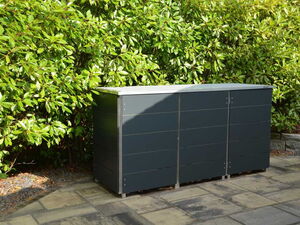 . The image depicts an outdoor setting with a large, dark-colored storage container or bin situated on a paved surface. Behind the container is a lush hedge or row of green leafy plants, providing a natural backdrop. The scene appears to be a garden or patio area.