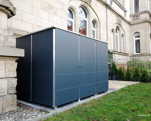 Ein modernes, dunkles Gartenhaus steht neben einem Steingebäude mit dekorativen Fenstern und umgeben von Grün.
