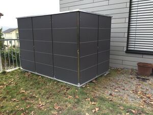 . The image shows a metal storage shed placed on a patch of grass next to a building. There are fallen leaves scattered on the grass, and a brown empty pot is visible near the building. The shed has smooth, dark-colored panels and is located near a metal railing.