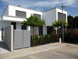 . The image depicts a modern residential building with flat, clean lines and minimalist design. The walls of the house are white, and the windows have horizontal shutters. There are small trees planted near the entrance, a paved walkway, and fencing along the perimeter. A streetlight stands in front of the house, and there is a sidewalk with some greenery.