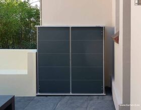 . The image shows a sleek, modern outdoor storage cabinet against a wall in a courtyard or garden area. It has a simple, flat design with smooth, dark gray panels. There is a patch of greenery in the background, suggesting the setting is part of a garden or patio.