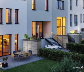 . This image shows a modern residential building with outdoor living spaces. The scene includes:

1. A patio area with a dining table and chairs.
2. Exterior lighting creating a cozy ambiance.
3. Several large windows showing lit interiors.
4. Landscaping with hedges and shrubs.
5. Stairs leading from a higher level down to the garden.

Overall, it depicts an elegant and well-maintained outdoor setting.
