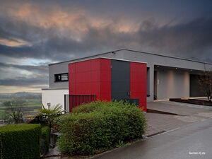 . Modernes Haus mit roter und grauer Fassade, üppigem Grün im Vordergrund, unter bewölktem Himmel.