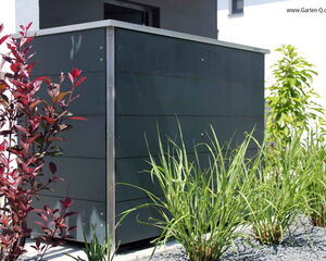 . The image shows a tidy outdoor area featuring a modern-looking structure, possibly a storage unit or enclosure, with sleek, dark paneling and metal trim. It is surrounded by various plants, including green grasses and a red-leafed shrub, set on a bed of dark gravel. The overall design is contemporary and minimalistic.