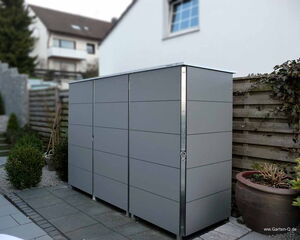 The image shows a sleek, modern storage cabinet or shed situated outdoors, likely in a garden or backyard. It is constructed with smooth, gray panels and has a lockable door. The surroundings include a paved area, a wooden fence, and some plants and shrubs, suggesting a tidy and organized outdoor space.