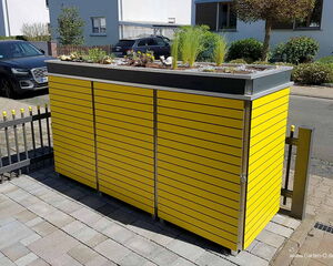 . The image shows a modern outdoor storage unit with a yellow slatted design. The top of the unit is a planter with various plants and decorative stones. It's located on a paved area, possibly a driveway, with cars and houses in the background.