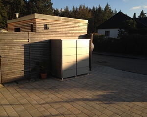 . The image shows an outdoor scene with a paved area. There's a modern-looking rectangular metal structure, possibly an outdoor storage unit, placed against a wooden wall or fence. A potted plant is also visible near the base of the wall. In the background, there are trees and a neighboring house with a sloped roof. The scene is bathed in sunlight, indicating that it might be late afternoon or early evening.