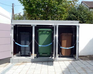 . The image shows a series of enclosed compartments containing three differently colored wheelie bins: black, green, and brown. These compartments appear to be part of an organized waste management system. The bins are secured with chains and are situated on a paved area, with trees and buildings visible in the background.