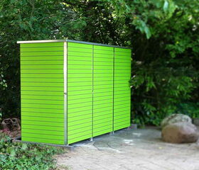 . The image shows a sleek, modern green shed situated outdoors amidst foliage. The shed has horizontal slats with a smooth surface, and it stands on a paved area surrounded by greenery, including trees and plants. Some large rocks are visible nearby on the ground.