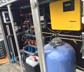 The image displays a complex plumbing system, possibly for a pool or spa. It includes:

1. Numerous PVC pipes and valves with orange handles.
2. A yellow electronic control unit labeled "dinotec".
3. A large blue tank, which could be a filter or water storage.
4. Various gauges and connectors.
5. Electrical wiring and switches.
6. A container with a red cap, possibly for chemicals or additives.

This setup appears to manage water flow and treatment.