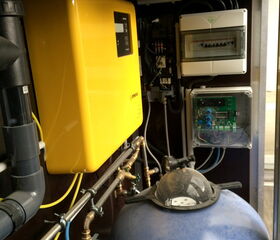 The image shows a compact utility area with the following components:

1. A large yellow box-like mechanism, likely an electronic device or inverter.
2. Several gray and black pipes, valves, and fittings, suggesting a plumbing or water filtration system.
3. A blue tank at the bottom, possibly a pressure tank or water filter.
4. Electrical panels and wiring on the wall, indicating a control system.
5. Various connectors and fittings, showing an integration of electrical and plumbing systems.

The setup appears to be part of a technical system, possibly related to water treatment or renewable energy.