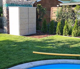 The image shows a garden scene with a small pool in the foreground. There is a lawn area with a yellow pool noodle lying on the grass. In the background, there is a modern storage unit, possibly for garden tools or equipment, and next to it, a stack of firewood secured by a metal frame. Some small trees and shrubs are planted along the edge of the garden, and a wooden shed is also visible behind the plants.