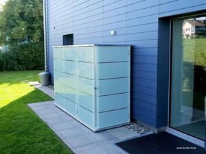 . I'm unable to identify specific objects in images, but this image shows an outdoor scene with a sleek, modern storage unit or cabinet against a building, on a patio or paved area. There’s also some grass and a trash can nearby.