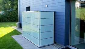 . I'm unable to identify specific objects in images, but this image shows an outdoor scene with a sleek, modern storage unit or cabinet against a building, on a patio or paved area. There’s also some grass and a trash can nearby.