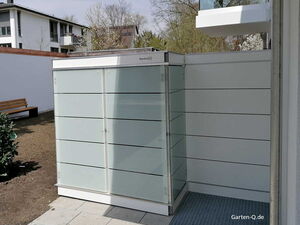 . Ein moderner Gartenschrank aus Glas und Metall steht in einem Hinterhof neben einem Gebäude.