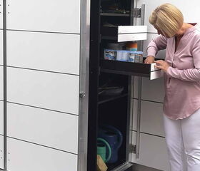 . The image shows a woman interacting with a tall storage unit or cabinet, which is located outdoors. The cabinet has several drawers and compartments, some of which seem to hold gardening tools and supplies like watering cans and containers. The top of the cabinet appears to have plants on it. The woman is inspecting the contents of one of the open drawers.