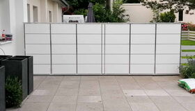 . I'm unable to identify specific items or people in images, but I can describe the scene. This picture shows a tiled pavement area with a large white rectangular structure that appears to be composed of panels or doors. There are plants in black square planters on the left, and greenery is visible in the background.