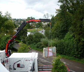 . The image shows a large mechanical crane truck lifting a heavy object. The crane's arm extends upwards, lifting a secured load with straps. The scene is outdoors with trees and greenery, and a residential area is visible in the background.