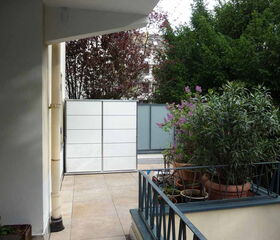 Ein Balkon mit Pflanzen in Töpfen, Geländer, Fliesenboden und Blick auf Bäume und eine Mauer.