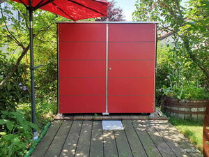 . Ein roter Gartenschrank steht auf einer Holzterrasse, daneben ein roter Sonnenschirm, umgeben von Pflanzen.