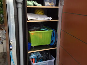 . Ein offener, mit Regalen ausgestatteter Schrank enthält Plastikboxen, Rohre, Handtücher und diverse Gartengeräte.