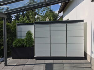 . Eine Terrasse mit Glasüberdachung, Sichtschutzwand und Blumenkübel mit grünem Strauch.