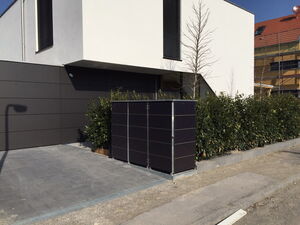 . The image depicts a modern building with a minimalist design. The building has contrasting light and dark panels with a smooth, sleek finish. There is a hedgerow along one side, partially obscuring a structure that resembles a small shed or utility box, possibly for solar or electrical equipment. The foreground includes a paved driveway and some landscaping with small trees.