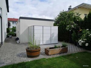 . The image shows an outdoor space with several features:

1. **Buildings**: There appears to be a white structure with a flat roof and another yellow building with a sloped roof in the background.
2. **Metal Structure**: A white, rectangular metal storage unit or cabinets.
3. **Planters**: There are two planters, one round wooden barrel and one rectangular wooden box, containing various plants.
4. **Pathway**: Paved with rectangular stones.
5. **Garden**: Includes various plants and bushes, some with flowers.
6. **Lawn**: A small grass area is visible in the foreground.

The setting looks like a residential backyard or garden area.