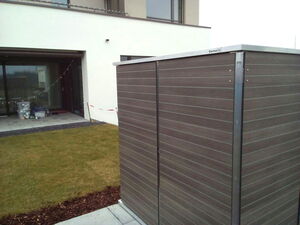. Ein Außenbereich mit Rasen, Terrasse und Gartenbox neben einem modernen Gebäude.