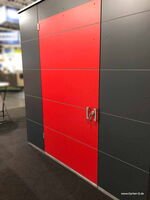 The image shows a structure with red and dark gray panels. It appears to be a modern storage unit or cabinet, possibly part of a modular garden shed system. There is a lock and handle on the center panel, suggesting it is a functional door. The structure is located indoors, suggesting it is on display, possibly at a trade show or exhibition. The website "www.Garten-Q.de" is visible in the corner.
