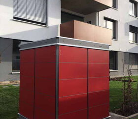 . The image shows a red shed-like structure placed in front of a modern apartment building. The shed has a metallic frame and is covered in red panels. The apartment building is contemporary, with multiple floors and balconies, and there is grass and paving surrounding the shed.