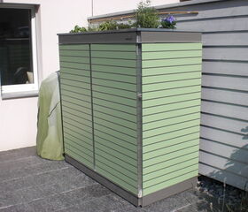 The image shows a sleek, green outdoor storage unit against a wall. The unit has horizontal slats and appears to be used for storage, likely for gardening tools or equipment. There is a cover beside it, possibly protecting another piece of garden equipment, and some plants can be seen on top of the storage unit, suggesting it might also serve as a planter. The setting is outside, on a tiled or paved area near a building with a window.