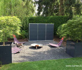 . The image shows a garden setup with two colorful lounge chairs on a gravel area, flanking a circular fire pit in the center. There are two large plant containers with greenery nearby, and a large storage unit in the background. The surrounding area is lush with trees and bushes.