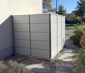 . The image shows a modern, rectangular outdoor storage unit or shed placed next to a building. The shed is made of metal panels and is situated on a gravel and stone path, surrounded by some greenery.