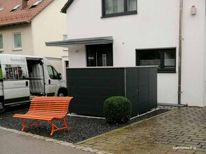 . The image shows the exterior of a house with a white wall and some windows. In front of the house, there's a modern black structure, possibly a storage unit, with a small neatly trimmed shrub next to it. Nearby, an orange bench is placed on a gravel surface. To the left, a parked van is partially visible. The setting appears to be a driveway or garden area.