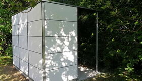 . Ein modernes Gartenhaus mit weißen Paneelen, umgeben von Bäumen im Schatten.