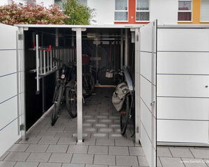 . The image shows a storage shed that contains at least two bicycles, a ladder, and some other tools or equipment. The shed is open, revealing its contents, and it is situated on a paved area. The door is open, allowing a clear view inside.