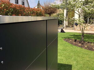 . The image shows an outdoor scene with a modern metal structure, possibly a cabinet or enclosure, in the foreground. In the background, there's a garden with green grass, a tree with white blossoms, and a house with a brown tiled roof. The setting is bright with sunlight and a clear blue sky.