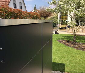 . The image shows an outdoor scene with a modern metal structure, possibly a cabinet or enclosure, in the foreground. In the background, there's a garden with green grass, a tree with white blossoms, and a house with a brown tiled roof. The setting is bright with sunlight and a clear blue sky.