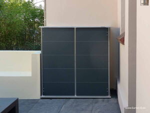 . The image shows a sleek, rectangular outdoor storage unit with a modern design. It has a dark-colored front and appears to have a flat top. The unit is situated against a wall on a patio or outdoor area. There's some greenery visible in the background. The website "www.Garten-Q.de" is written in the bottom right corner of the image.