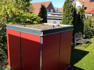 . The image shows the following elements in an array:

1. **Red Garden Shed**: A modular garden shed with a red exterior and a flat, green roof.
2. **Green Roof**: The roof is planted with various small plants, likely succulents or similar vegetation.
3. **Garden Bench**: An ornate metal bench next to the shed.
4. **Trees and Shrubs**: Various types of trees and shrubs surrounding the area, providing greenery.
5. **Lawn**: A well-maintained grass lawn.
6. **Houses**: Residential buildings with red-tiled roofs in the background.
7. **Paved Pathway**: A pathway made of stones or bricks leading through the garden.

The scene is a backyard or garden space that appears tidy and well-kept.
