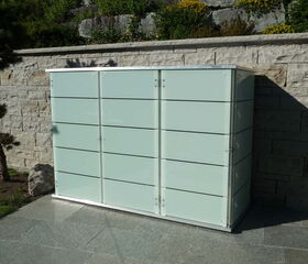 . The image shows a modern, sleek outdoor storage cabinet or shed placed against a stone wall. It has a smooth, glossy finish with horizontal lines and appears to be made of metal or another durable material. It's situated in a landscaped outdoor area with trees, shrubs, and a tiled ground surface.