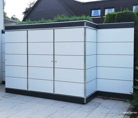 . The image depicts a modern outdoor storage unit or shed. It has a sleek design with clean, white panels and a flat roof. The storage unit is situated on a tiled patio, with some greenery visible in the background.