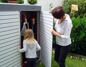 . The image depicts two people, possibly an adult and a child, standing in front of an outdoor storage cabinet or garden shed. The adult is opening the doors of the shed while the child is looking inside. Inside the shed, various garden tools can be seen, including a rake and a broom. The setting appears to be a garden or backyard with green foliage around.